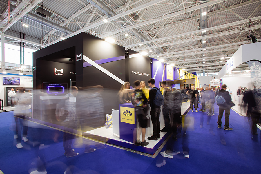 Vista laterale dello stand Magneti Marelli Parts & Services ad Autopromotec 2025: in primo piano il tunnel Marelli con tecnologie di frontiera, sullo sfondo il logo aziendale giallo e blu e visitatori nel corridoio adiacente.